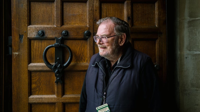 Male volunteer in glasses and a gilet, wearing a black volunteer lanyard and a name badge. He is waiting to greet people coming into the manor house at Sizergh, and stands in front of the large, dark oak door with it's black door knocker.